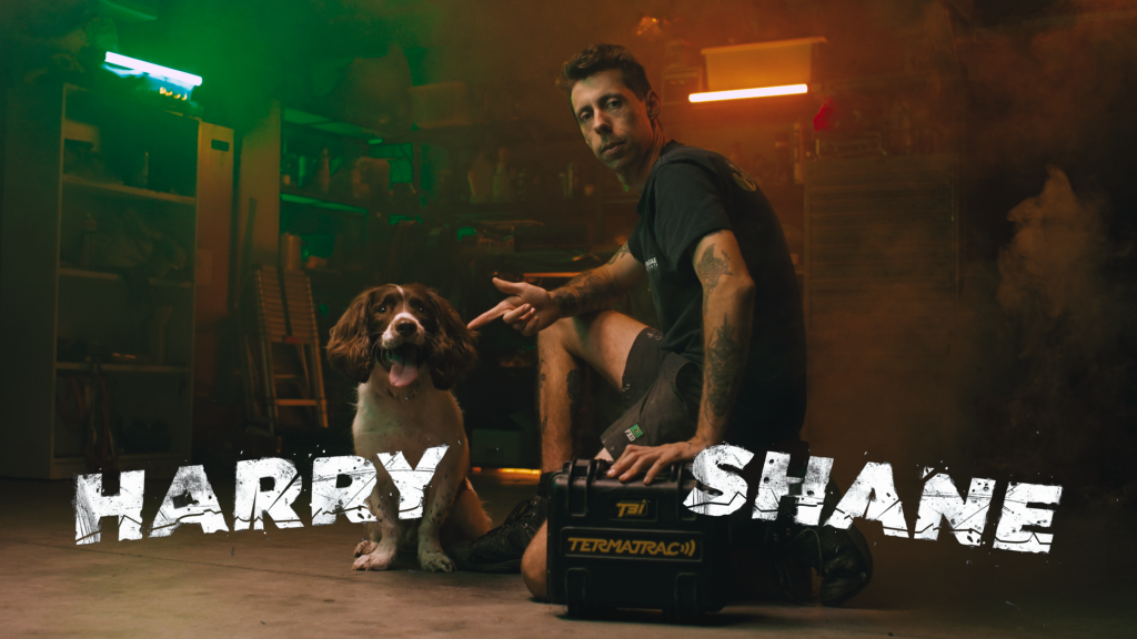 A man named Shane, kneeling beside a dog named Harry, holding a Termatrac device in a dimly lit workshop.