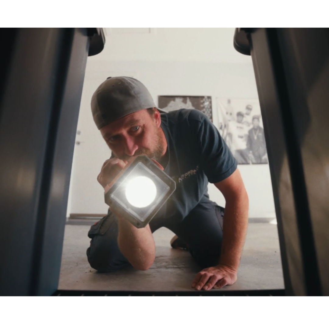 A man wearing a backwards cap and a dark shirt, holding a flashlight, is kneeling on the ground and looking into a narrow space between two objects.